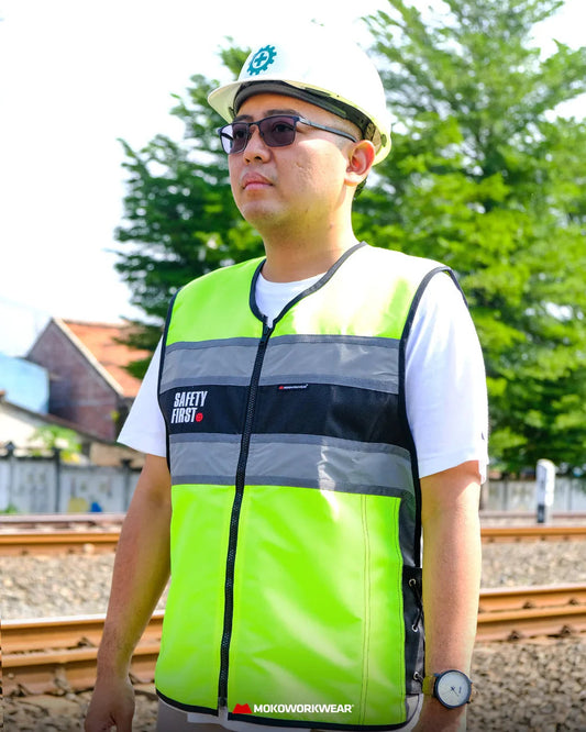 Man wearing safety vest and hard hat standing near railway tracks with trees and buildings in background