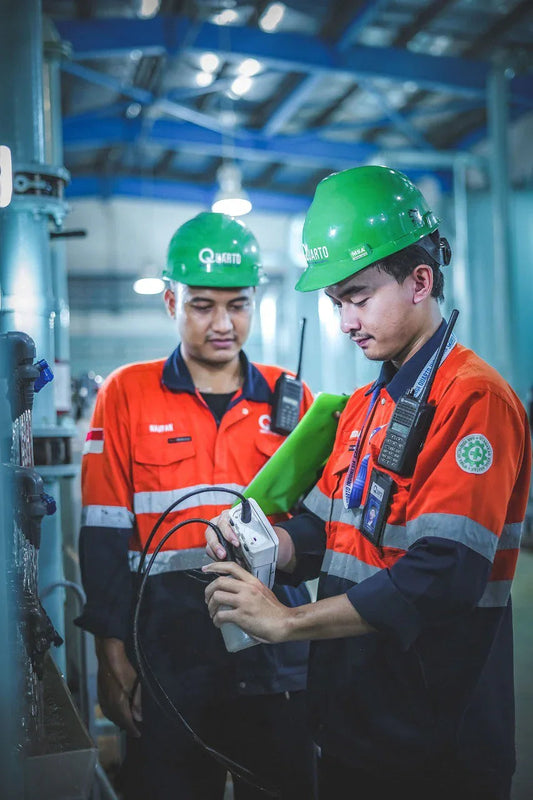 Two industrial workers in green helmets and orange safety uniforms using a measuring device in a factory setting