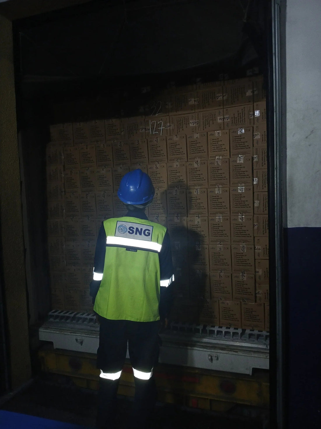 Worker in reflective safety vest and blue helmet inspecting stacked boxes in dimly lit warehouse