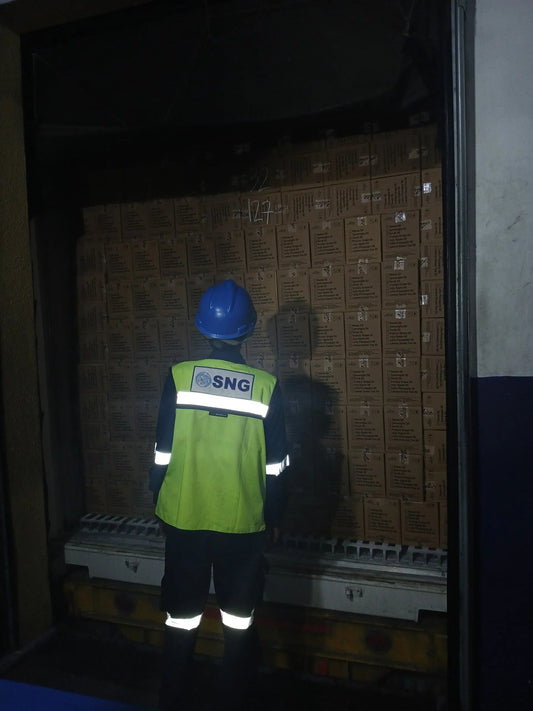 Worker in reflective safety vest and blue helmet inspecting stacked boxes in dimly lit warehouse